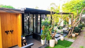 A modern outdoor seating area with a wooden door, potted plants, and a black structure with glass windows. - Baanpoon Apartment (Koh Samui)