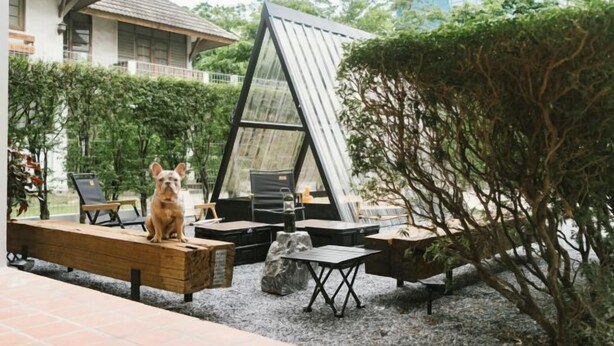 A modern outdoor seating area with a dog, a glass structure, and a building in the background. - My Sukhumvit Villa (Bangkok)