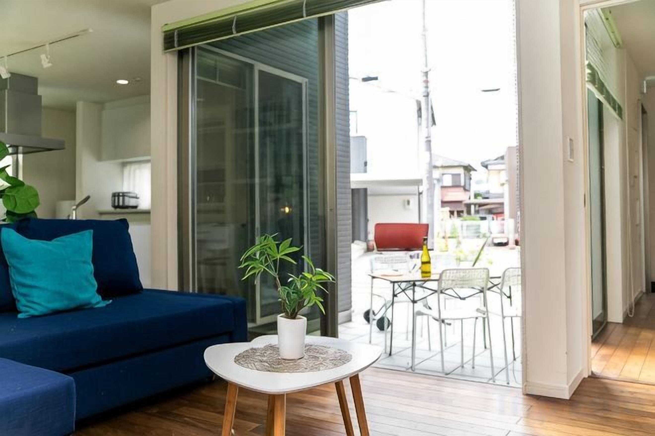 Una sala de estar moderna con un sofá, una mesita con una planta y un ventanal corredizo que da a un espacio exterior con una mesa y sillas.
