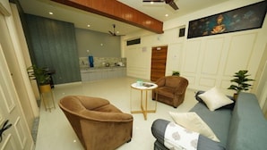 Una sala de estar con un sofá, sillones y una mesa de centro. Se observa una cocina al fondo con armarios y una encimera.
