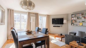 A modern living room with a wooden dining table, two chairs, a TV mounted on the wall, and a bookshelf. - La Maison de Pierre et Valérie Saint Catherine (Honfleur)