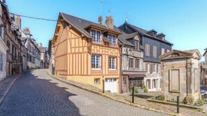 A cobblestone street lined with traditional half-timbered buildings, a small stone structure, and a clear blue sky. - La Maison de Pierre et Valérie Saint Catherine (Honfleur)