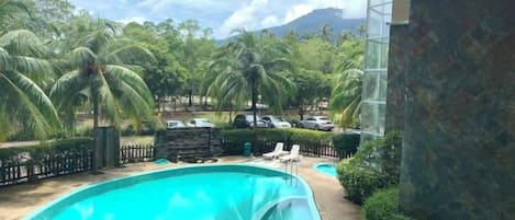 A swimming pool with lounge chairs, surrounded by palm trees and a mountain in the background.
