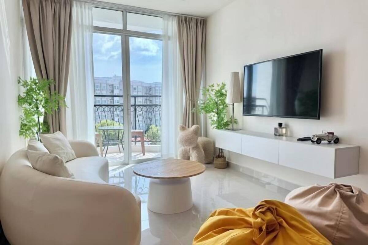 A modern living room with a sofa, a coffee table, a flat-screen TV, and a balcony with a view of buildings.