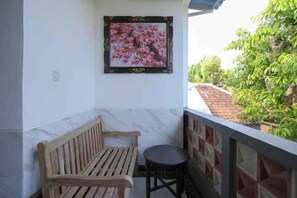 A balcony with a wooden bench and a small round table, a framed picture on the wall, and a view of a garden. - RedDoorz @ Lotus House Seminyak (Bali)