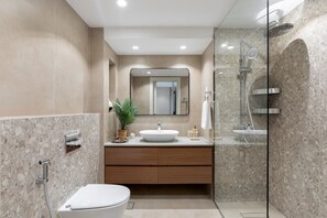 A modern bathroom with a freestanding toilet, a sink with a vessel basin, a mirror above the sink, a shower with a glass enclosure, and a potted plant on the vanity. - Chic Downtown Living w Dubai Cityscape Views (Dubai)