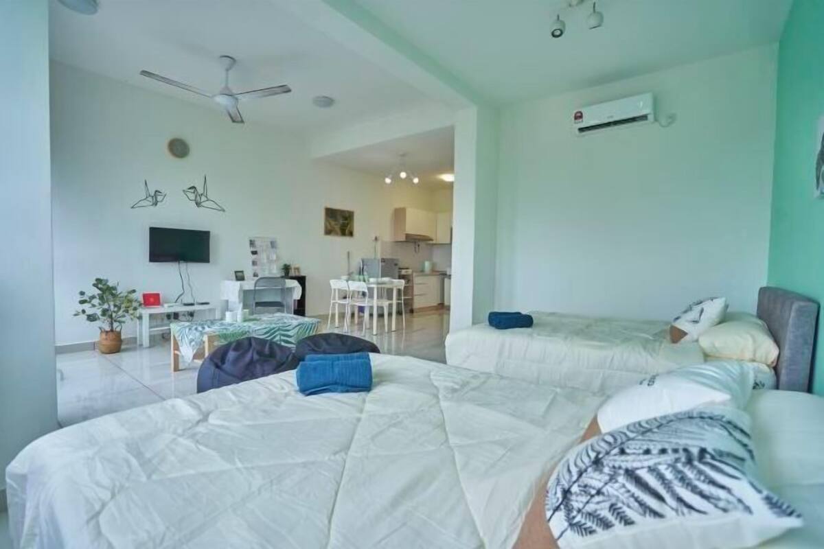Une chambre d’hôtel avec deux lits, une télévision, un ventilateur de plafond et, au fond, une kitchenette.