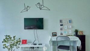 A room with a TV mounted on the wall, a white desk with a chair, a plant, and a bulletin board.