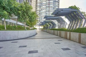 Un sendero urbano moderno con un paseo curvo, bordeado de follaje y un edificio alto al fondo.