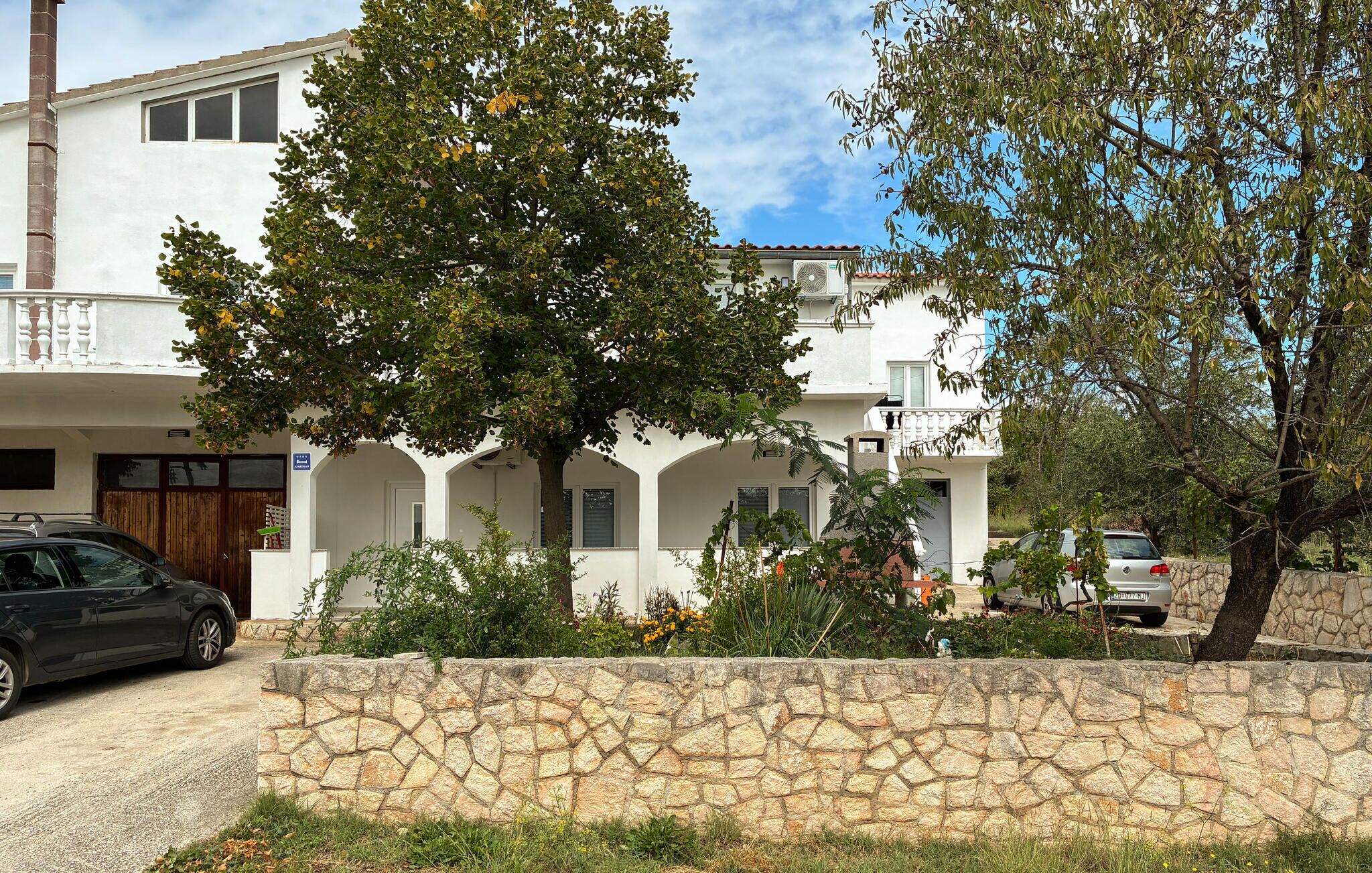 A two-story building with a stone wall in front, a parked car, and a tree with yellow flowers.