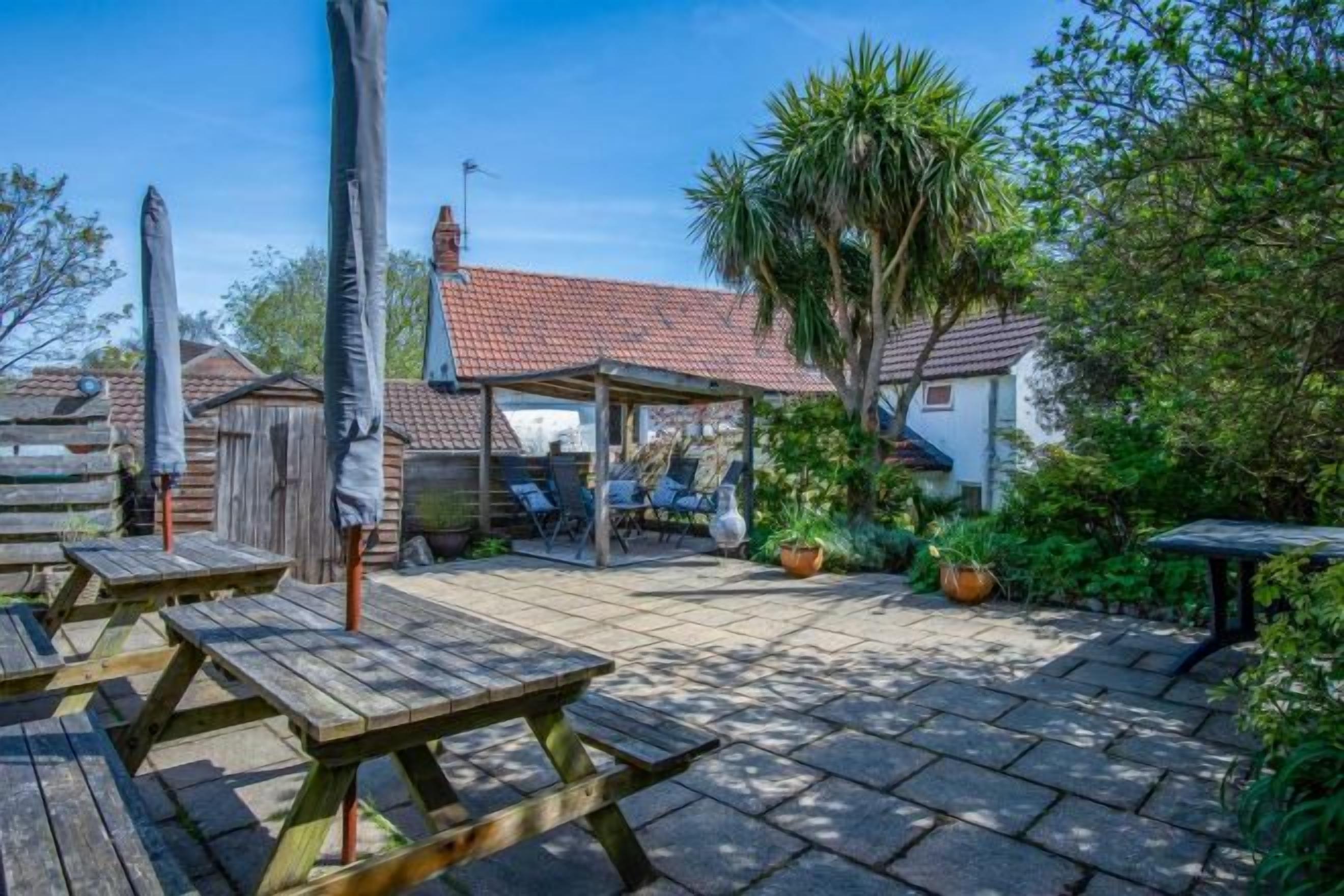 A patio area with wooden tables and umbrellas, surrounded by potted plants and a house with a tiled roof.