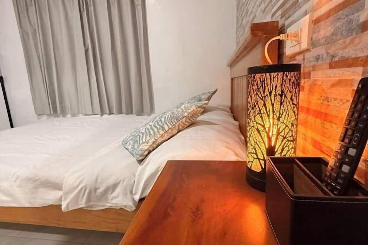 A neatly made bed with white linens and a patterned pillow, a wooden nightstand with a lamp, and a brick wall with a window and curtains.