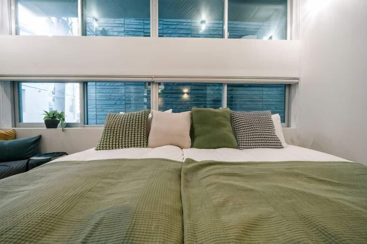 Un lit recouvert d’une couverture verte, une fenêtre donnant sur la ville et une plante posée sur le rebord de la fenêtre.