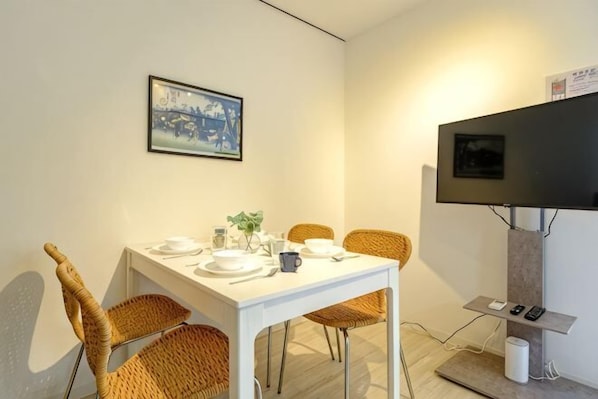 Un comedor con una mesa lista para cuatro personas, rodeada de sillas de mimbre, un televisor montado en la pared y un cuadro enmarcado sobre la mesa.
