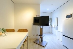 Una habitación de hotel moderna con un televisor de pantalla plana montado sobre un soporte, una mesa de comedor con sillas, un refrigerador y una ventana con cortinas.