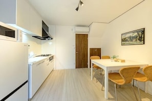 Une cuisine de taille réduite avec des appareils électroménagers blancs, un coin repas avec des chaises en osier et un tableau encadré au mur.