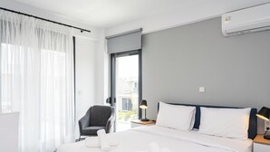 Ein Hotelzimmer mit einem Bett, einem Sessel, einem Nachttisch, einer Klimaanlage und einem Fenster mit durchsichtigen Vorhängen.