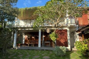A two-story building with a covered porch, a brick wall, and a garden area with a tree and potted plants. - Munay Villa (Bali)