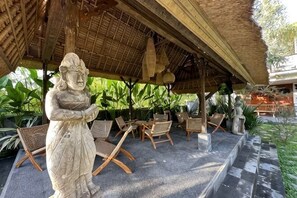 A covered outdoor seating area with stone statues, wicker furniture, and a thatched roof. - Munay Villa (Bali)