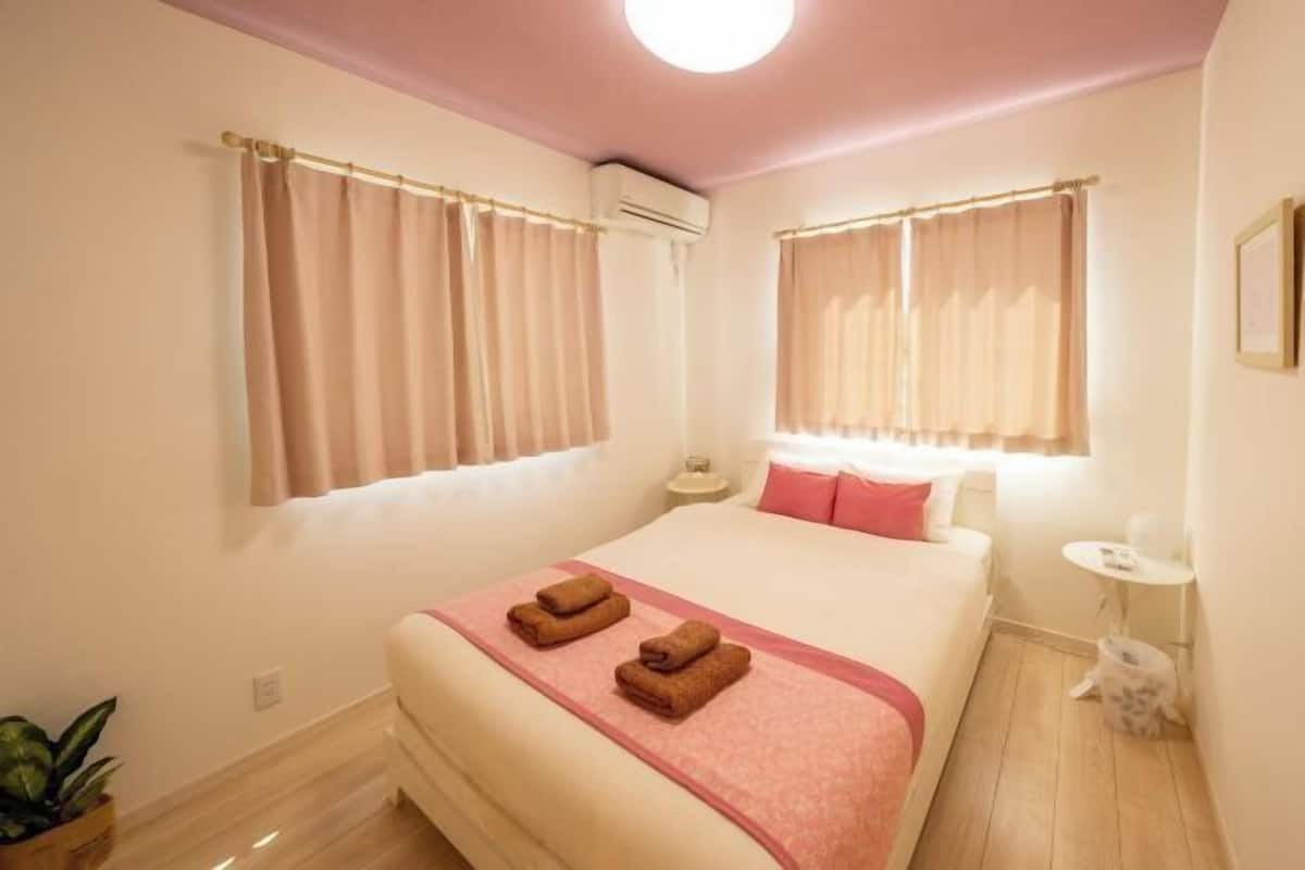A hotel room with a bed, two windows with curtains, a bedside table, a small round table, and a potted plant.