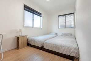 Ein kleines, sauberes Zimmer mit zwei Betten, einem Nachttisch, einer Lampe und einem Fenster.