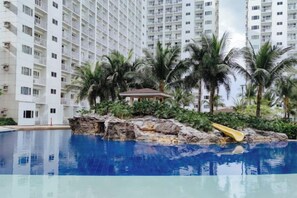 A swimming pool with a rocky island, palm trees, and a building in the background. - Lascianos SMDC Shore Residences by RedDoorz (Pasay)