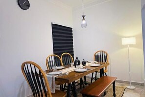 A dining area with a wooden table set for four, flanked by wooden chairs with black cushions. A black and white striped window blind is visible, and a floor lamp is on the right. - Wawasan Puchong Family-Friendly Spacious Home (Puchong)