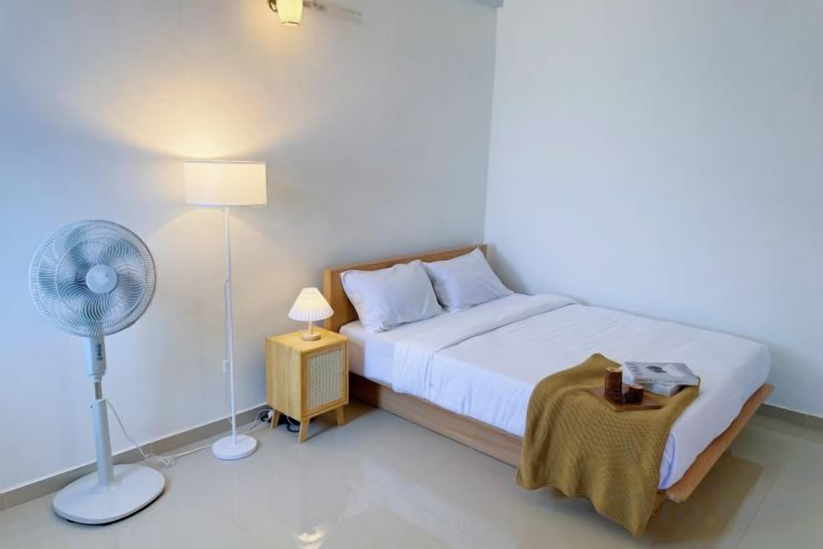 A bedroom with a bed, bedside table, lamp, and floor fan.