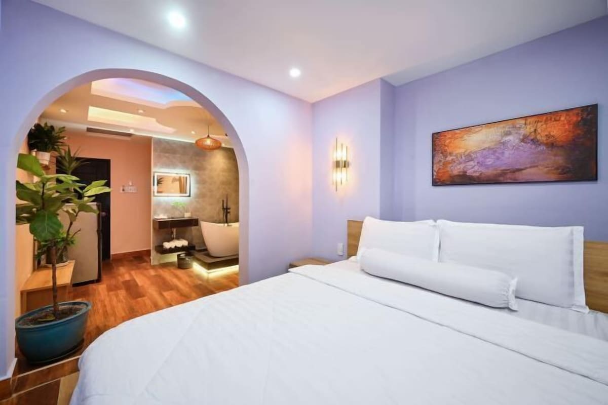 A modern hotel room with a large bed, a painting on the wall, and a bathroom visible through an archway.