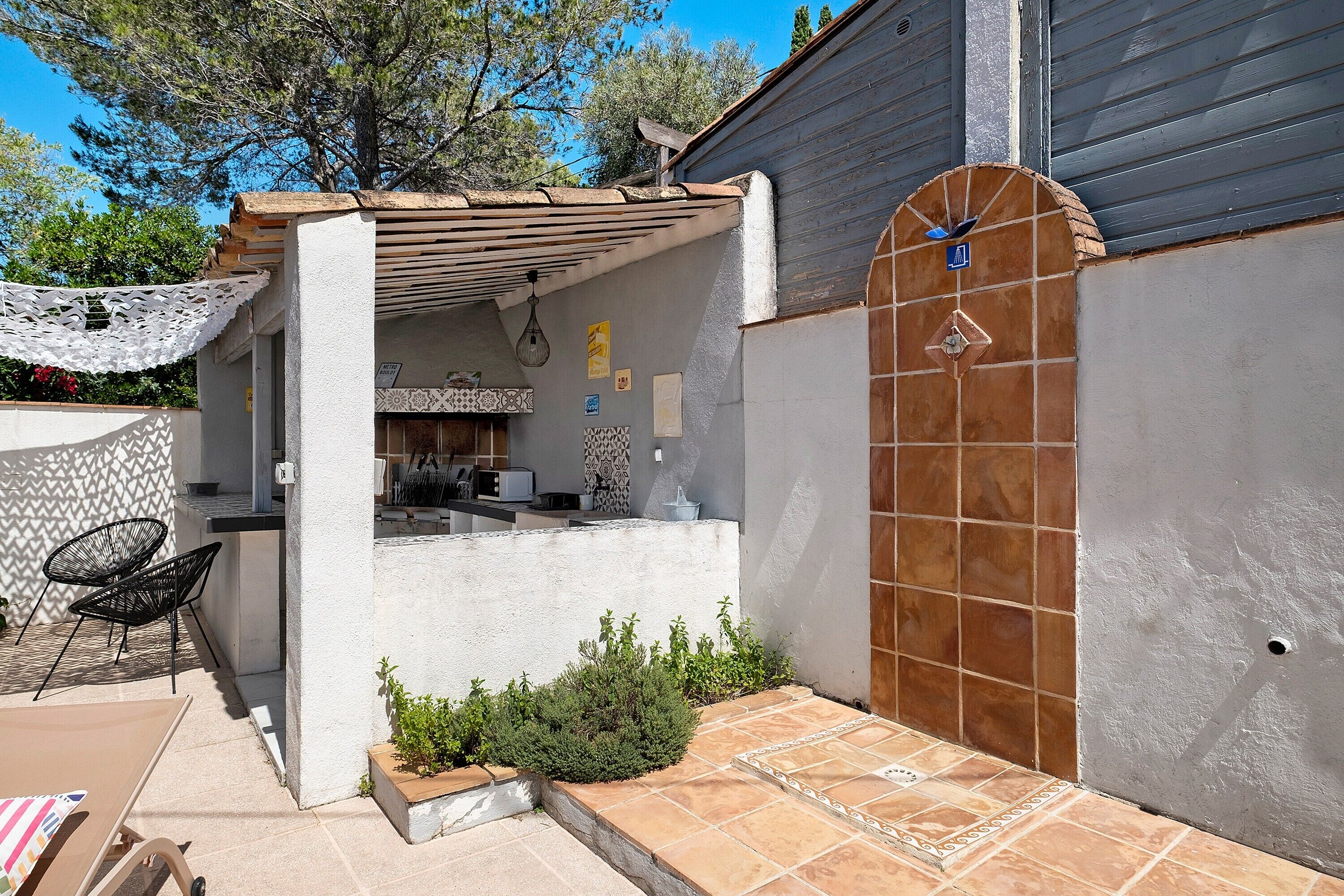 Un espacio de estar al aire libre cubierto con una pérgola, una cocina visible a través de una pared abierta y un patio con baldosas.