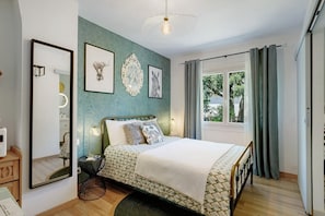 A bedroom with a bed, a mirror, framed artwork, a bedside table, and a window with curtains.