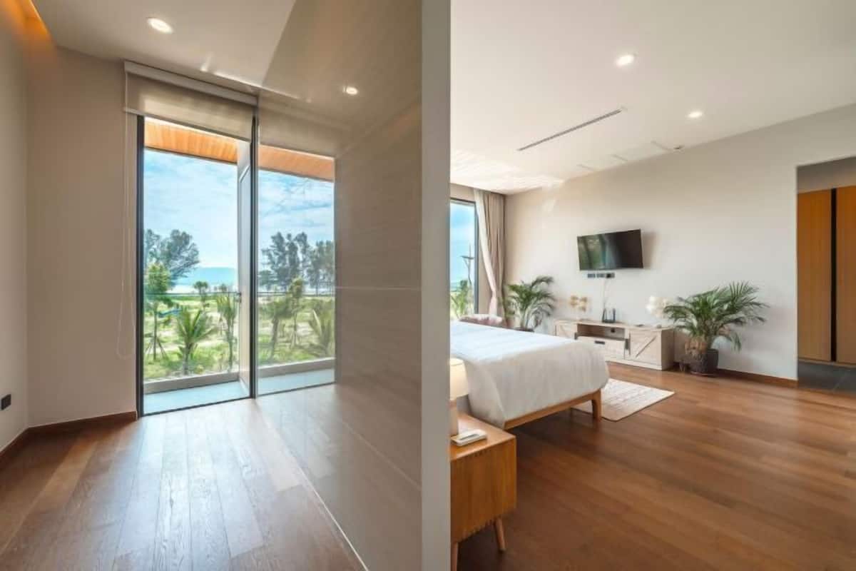 A modern bedroom with a large bed, a TV, and a balcony with a view of greenery.
