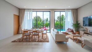 A spacious living room with a dining area, large windows, and a view of greenery. - Long One A5 (Phuket)