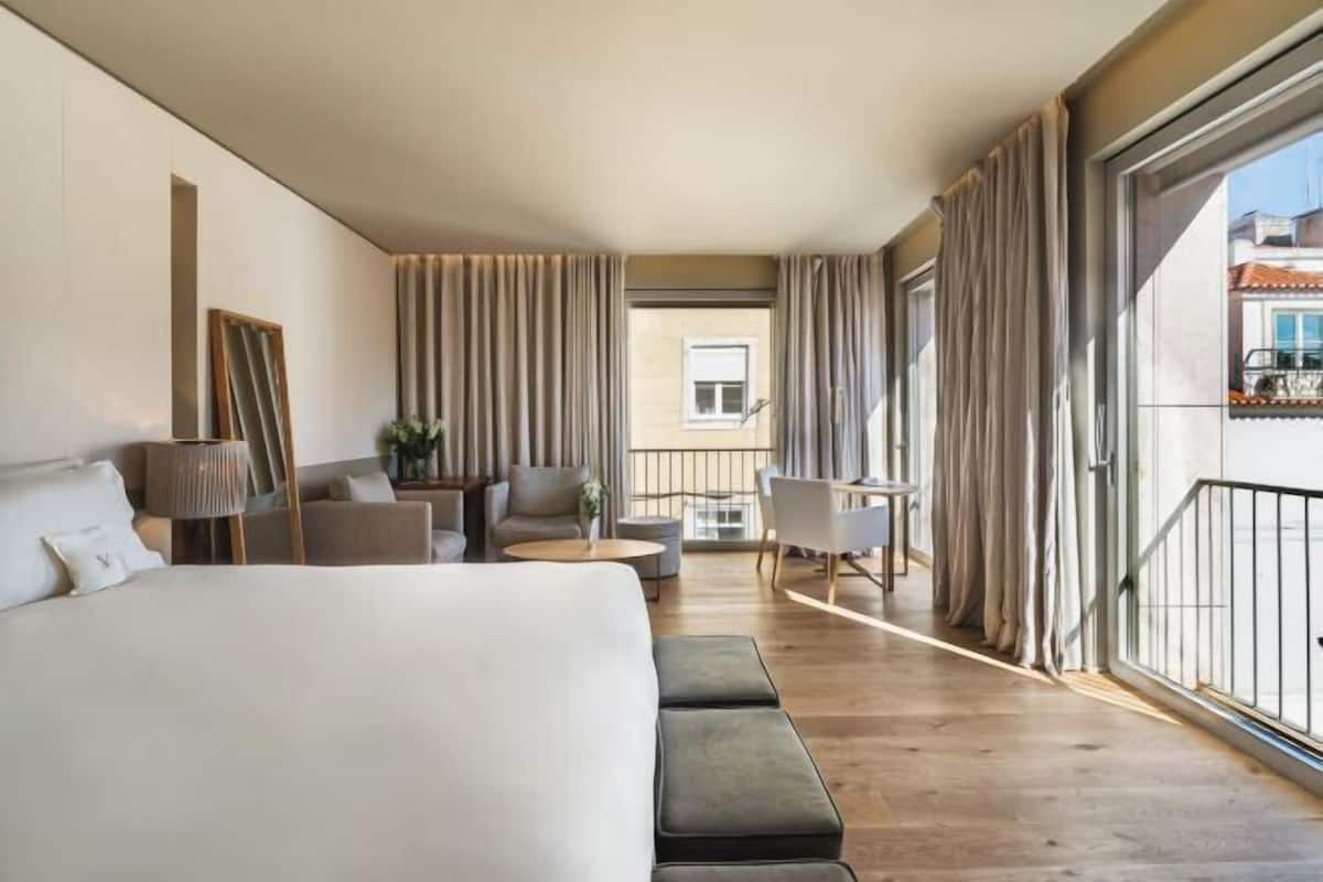 Habitación de hotel con una cama grande, una zona de estar con una mesa y sillas, y un balcón con cortinas.