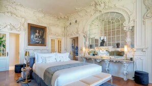 A luxurious bedroom with ornate white walls, a large bed with a bench, a mirror, and a painting on the wall. - Verride Palácio Santa Catarina (Lisbon)