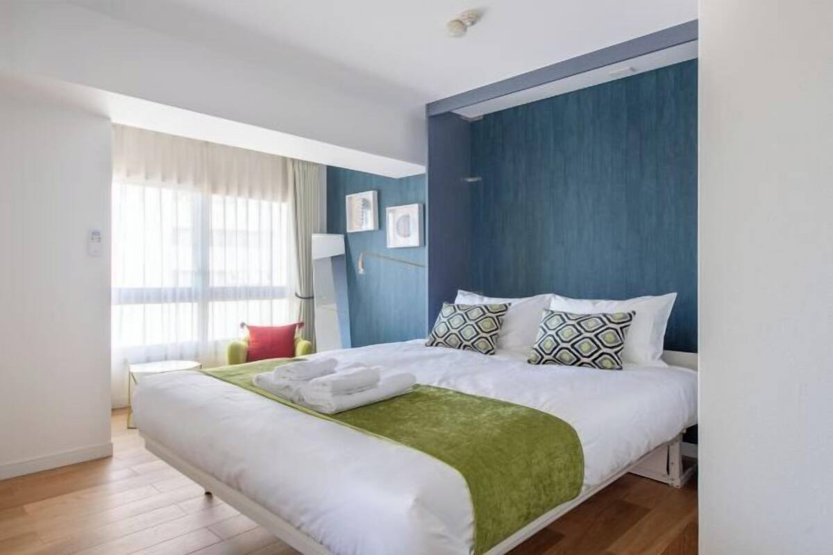 Una habitación de hotel con cama, una ventana con cortinas, una mesita de noche y un cuadro en la pared.