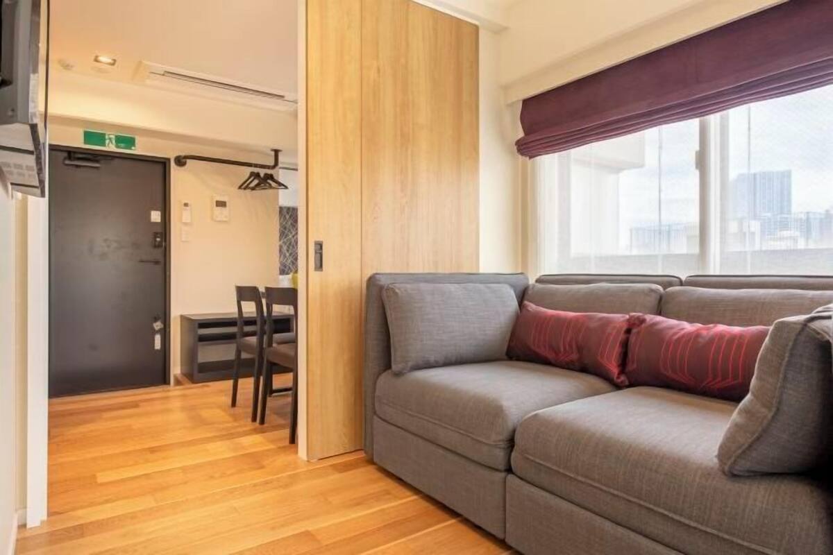 Una habitación moderna con un sofá gris, un separador de madera, un rincón comedor con sillas y una ventana con vistas.
