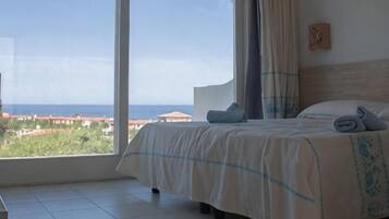 A bedroom with a large window, a bed with a blue and white bedspread, and a view of the sea.