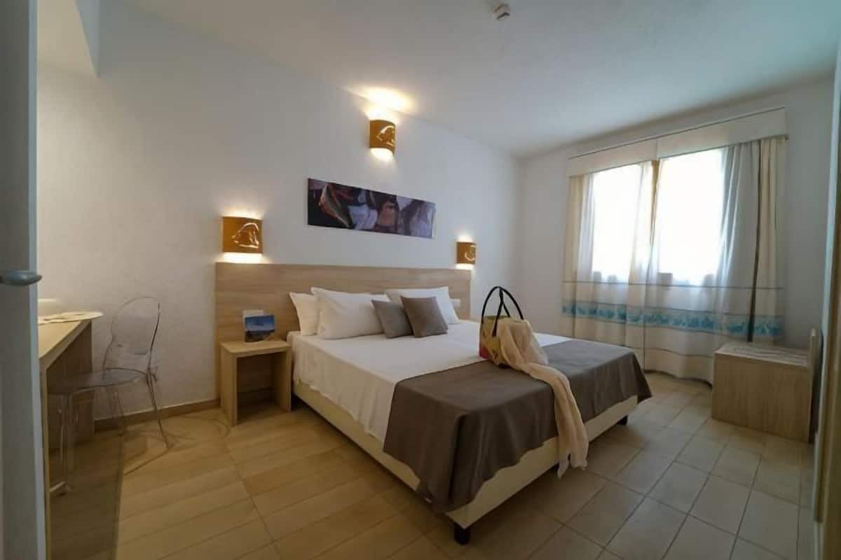 Una habitación de hotel con cama, mesitas de noche, escritorio, silla y ventana con cortinas.