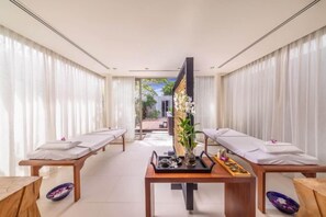 A spa room with two massage tables, a wooden table with a vase of flowers, and a view of the outdoors.