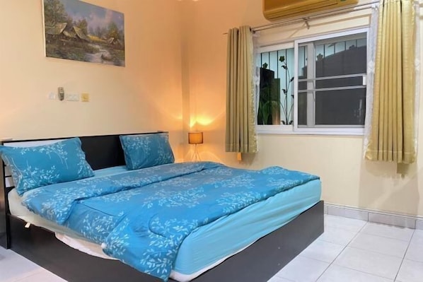 A bedroom with a bed, a nightstand, a lamp, a window with curtains, and a painting on the wall. - Royal park village (Pattaya)