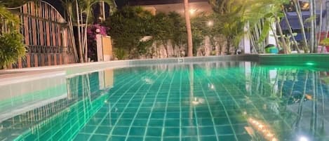 A swimming pool with a tiled surface, surrounded by palm trees and a building with a curved roof.