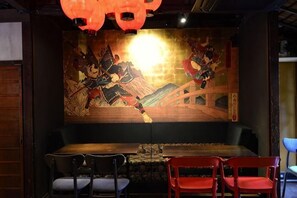 En restaurantindretning med et langt bord, røde og sorte stole og et muralmaleri på væggen.