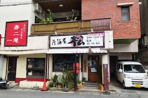 A building with a storefront featuring a sign with Chinese characters, a balcony with plants, and a white van parked in front. - Guest House Matsuyama (Naha)