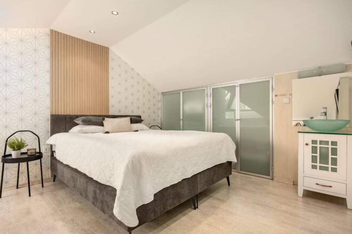 Un dormitorio con cama, mesita de noche, espejo, puerta corredera de vidrio y baño con lavabo y lavatrastos.