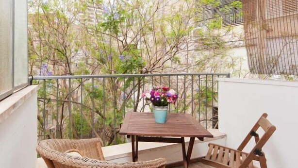 A balcony with wicker furniture, a small table with a vase of flowers, and a view of trees and buildings. - Stylish Apt w/ Charming Balcony in the Very Center (Tel Aviv)