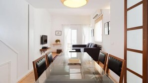 A modern living room with a glass dining table, black leather chairs, a sofa, a television, and a hanging light fixture. - Stylish Apt w/ Charming Balcony in the Very Center (Tel Aviv)