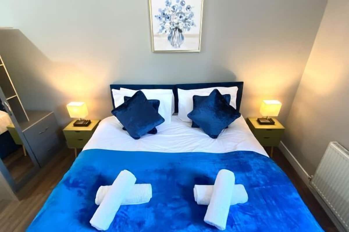 Ein Bett mit blauer Bettdecke und weißen Handtüchern, zwei dunkelblaue Kissen und ein gerahmtes Blumenbild über dem Bett.