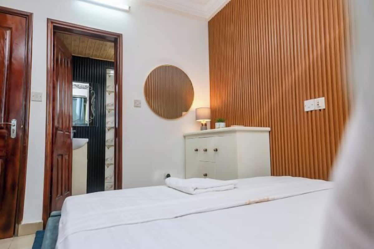 Une chambre d’hôtel avec un lit, une table de chevet avec une lampe, un mur recouvert de panneaux en bois et une porte donnant sur une autre pièce.