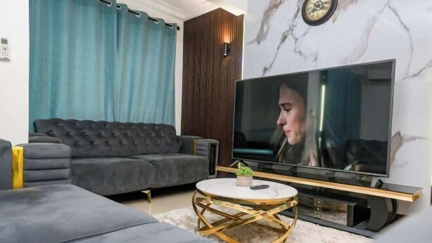A modern living room with a grey sofa, a round coffee table, and a flat-screen TV mounted on a marble wall. - T-Square Residence Makepe (Douala)